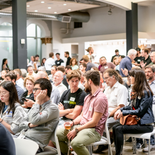 Ganz viele Menschen, sitzen auf Stühlen und hören zu.